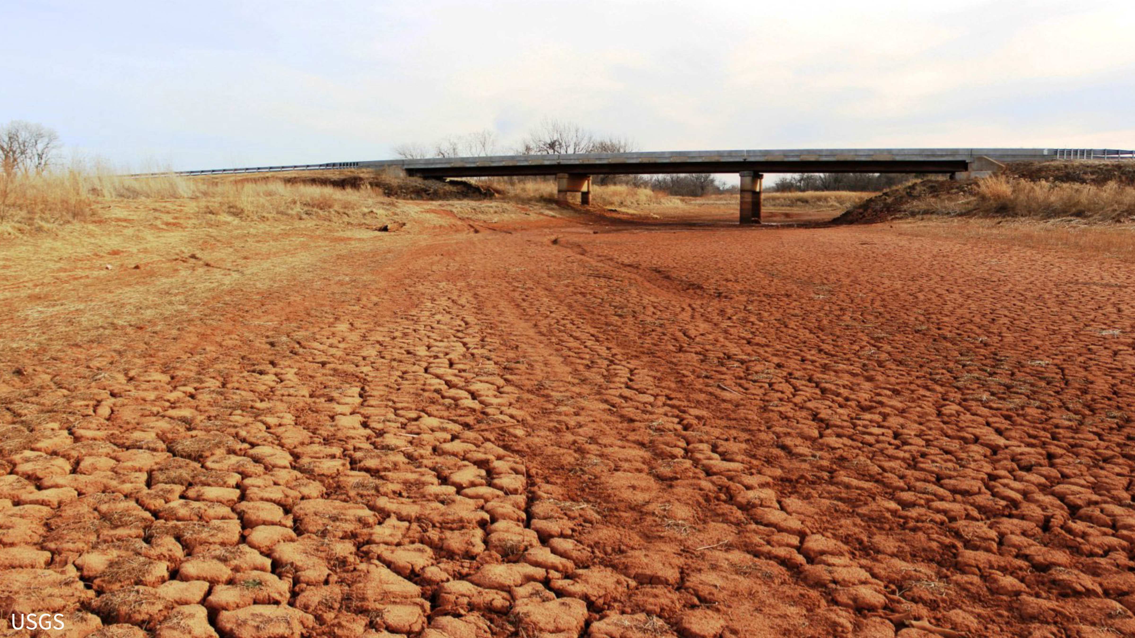 red clay based dirt cracked deeply from drought where a river once was. An over pass in the distance. Photo by USGS