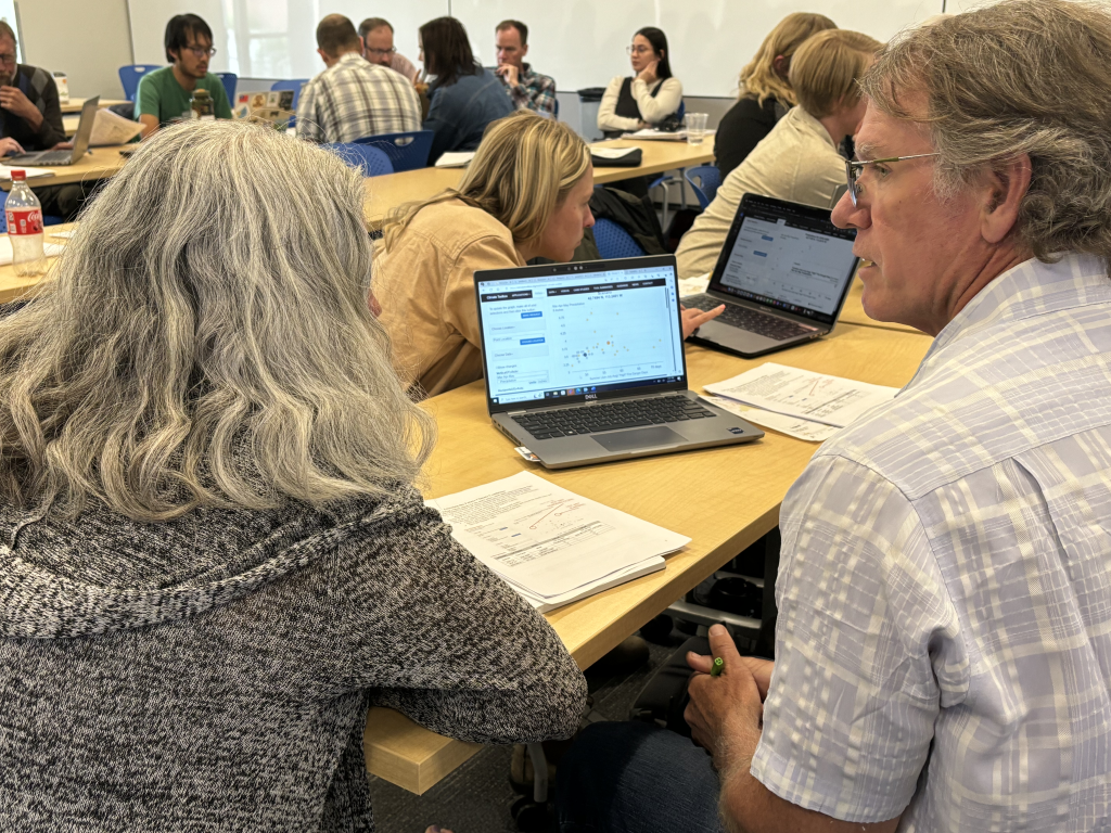 Two people discuss an activity with a laptop and paper in front of them. Other workshop attendees are in the background